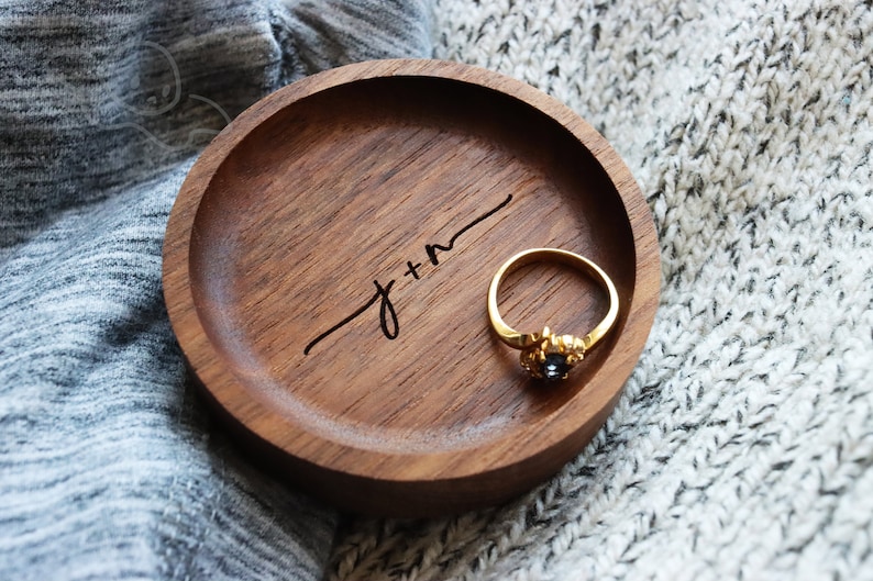 May include: A wooden ring dish with the inscription "J+M" and a gold ring with a black stone in the center.