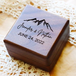 May include: A square dark brown wooden ring box with a smooth finish. The lid features a laser-engraved mountain range graphic above the names Jennifer & Nathan and the date JUNE 24, 2022, all centered in black text on the top surface.