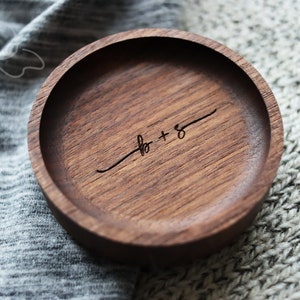 May include: A round wooden ring dish with a dark brown wood grain. The dish has the words "to + &infin;" engraved on the inside.