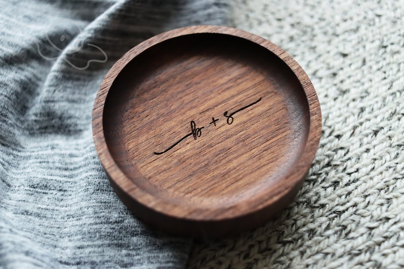 May include: A round wooden ring dish with a dark brown wood grain. The dish has the words "to + &infin;" engraved on the inside.