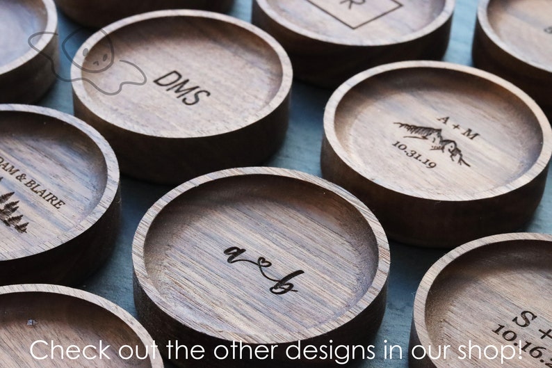 May include: A collection of wooden ring dishes with various engraved designs, including initials, dates, and a mountain range. The text "Check out the other designs in our shop!" is visible at the bottom of the image.