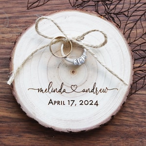 May include: A circular wooden slice ring bearer pillow featuring a natural bark edge and a light-colored center. The wood is engraved with the names Melinda and Andrew, a heart symbol, and the date April 17, 2024. Two rings are tied with twine.