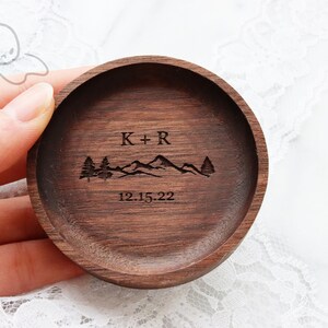 May include: A round wooden ring dish with a mountain range and two trees engraved on it. The dish is personalised with the initials "K+R" and the date "12.15.22".