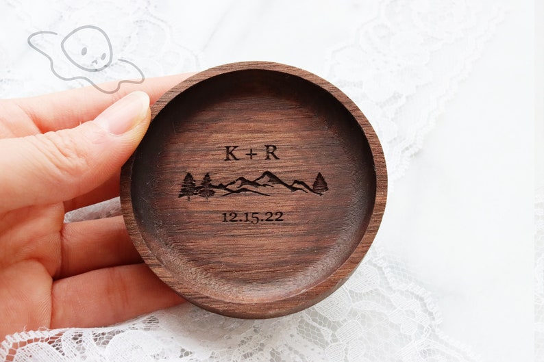 May include: A round wooden ring dish with a mountain range and two trees engraved on it. The dish is personalised with the initials "K+R" and the date "12.15.22".
