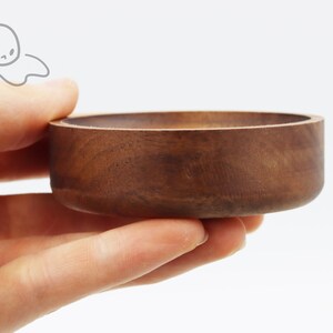 May include: A small, round, wooden bowl with a dark brown finish. The bowl is being held in a person's hand.