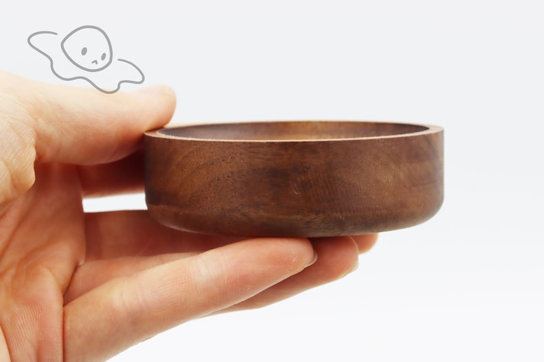 May include: A small, round, wooden bowl with a dark brown finish. The bowl is being held in a person's hand.