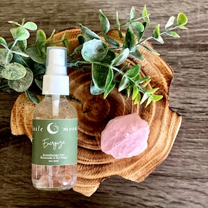 Op de afbeelding: Een doorzichtige glazen fles met een witte spuitdop en een groen label met de tekst "little moon Energize Aromatherapy Mist Handmade in San Diego 2oz | 60ml". De fles staat op een houten onderzetter met een roze rozenkwartskristal.