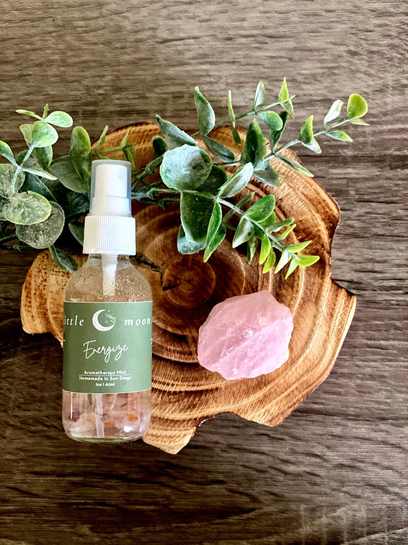 Op de afbeelding: Een doorzichtige glazen fles met een witte spuitdop en een groen label met de tekst "little moon Energize Aromatherapy Mist Handmade in San Diego 2oz | 60ml". De fles staat op een houten onderzetter met een roze rozenkwartskristal.