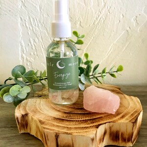 Op de afbeelding: Een doorzichtige glazen fles met een witte spuitdop en een groen label met de tekst "Little Moon Energize Aromatherapy Mist Handmade in San Diego 2oz | 60ml". De fles staat op een houten schijf met een roze rozenkwartskristal.
