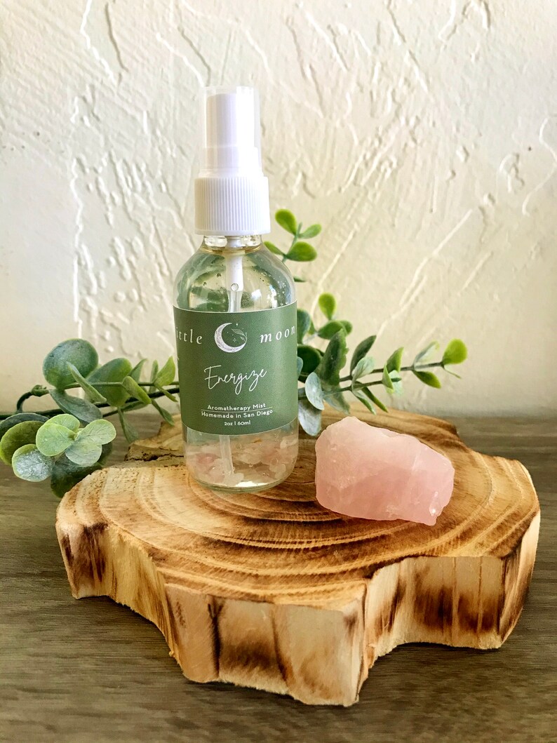 Op de afbeelding: Een doorzichtige glazen fles met een witte spuitdop en een groen label met de tekst "Little Moon Energize Aromatherapy Mist Handmade in San Diego 2oz | 60ml". De fles staat op een houten schijf met een roze rozenkwartskristal.