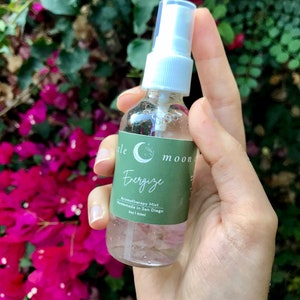 Op de afbeelding: Een doorzichtige glazen fles met een witte spuitdop en een groen label met de tekst "Little Moon Energize Aromatherapy Mist Handmade in San Diego 2oz | 60ml".