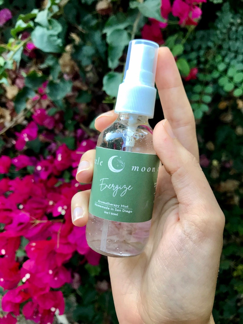Op de afbeelding: Een doorzichtige glazen fles met een witte spuitdop en een groen label met de tekst "Little Moon Energize Aromatherapy Mist Handmade in San Diego 2oz | 60ml".