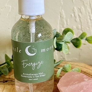 Op de afbeelding: Een doorzichtige glazen fles met een witte spuitdop en een groen label met de tekst "Little Moon Energize Aromatherapy Mist Homemade in San Diego 2oz | 60ml".