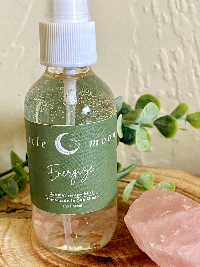 Op de afbeelding: Een doorzichtige glazen fles met een witte spuitdop en een groen label met de tekst "Little Moon Energize Aromatherapy Mist Homemade in San Diego 2oz | 60ml".