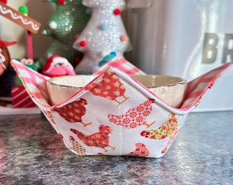 May include: A white bowl cozy with a red and white chicken pattern, holding a beige bowl. The cozy has a red and white gingham lining. Christmas decorations are in the background.