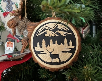 May include: A wooden Christmas ornament with a black silhouette of a mountain range, a forest, and a deer. The ornament is round and has a rustic wood grain texture.