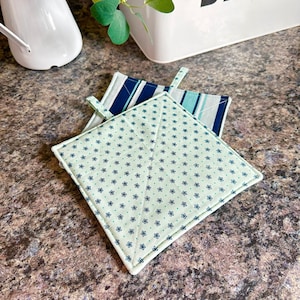 May include: Two green and blue patterned pot holders. One pot holder has a green background with small white flowers and the other has blue and white stripes.