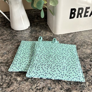 May include: Two green and blue patterned pot holders with a white background and a black floral design. The pot holders have loops for hanging.