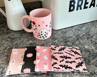 May include: A pink ceramic mug with a ghost and floral pattern sits next to four square fabric coasters. The coasters feature ghost, pumpkin, and bat patterns in pink and black. A white bread box is in the background.