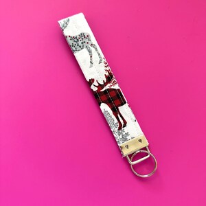 May include: White fabric key chain with a red and black plaid moose design. The key chain has a silver key ring.