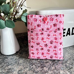 May include: A pink fabric book sleeve with a pattern of paw prints and bones. The sleeve has a pocket and is secured with a pink button and loop closure. The background includes a white vase with greenery and a white bread box.