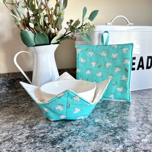 May include: A white ceramic bowl with a teal and white chicken print bowl cozy. The bowl cozy has a white interior and a teal exterior with a white chicken print. The bowl cozy is sitting on a gray countertop.