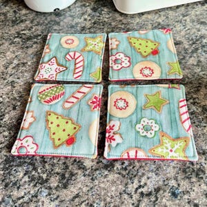 K&ouml;nnte beinhalten: Set aus vier quadratischen Stoffuntersetzern mit hellblauem Hintergrund und einem festlichen Weihnachtspl&auml;tzchen-Design. Jeder Untersetzer zeigt Lebkuchen in Rot, Gr&uuml;n und Wei&szlig; mit einem roten Rand.