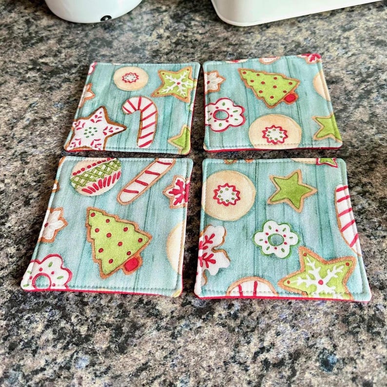 K&ouml;nnte beinhalten: Set aus vier quadratischen Stoffuntersetzern mit hellblauem Hintergrund und einem festlichen Weihnachtspl&auml;tzchen-Design. Jeder Untersetzer zeigt Lebkuchen in Rot, Gr&uuml;n und Wei&szlig; mit einem roten Rand.