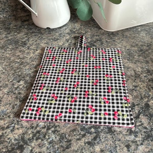 May include: Black and white checkered hot pad with a red cherry pattern and a pink binding. The hot pad has a loop handle.