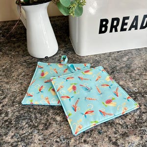 May include: Two square, light blue pot holders with a repeating pattern of colorful fishing lures. Each pot holder has a small loop for hanging. The pot holders are on a speckled countertop.