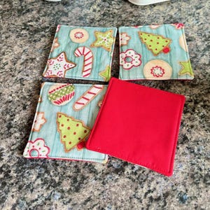 K&ouml;nnte beinhalten: Set aus vier quadratischen Untersetzern zum Thema Weihnachten. Drei Untersetzer haben einen hellblauen Hintergrund mit Lebkuchen-Designs in Rot, Gr&uuml;n und Wei&szlig;. Ein Untersetzer ist einfarbig rot. Die Untersetzer sind wahrscheinlich aus Stoff.