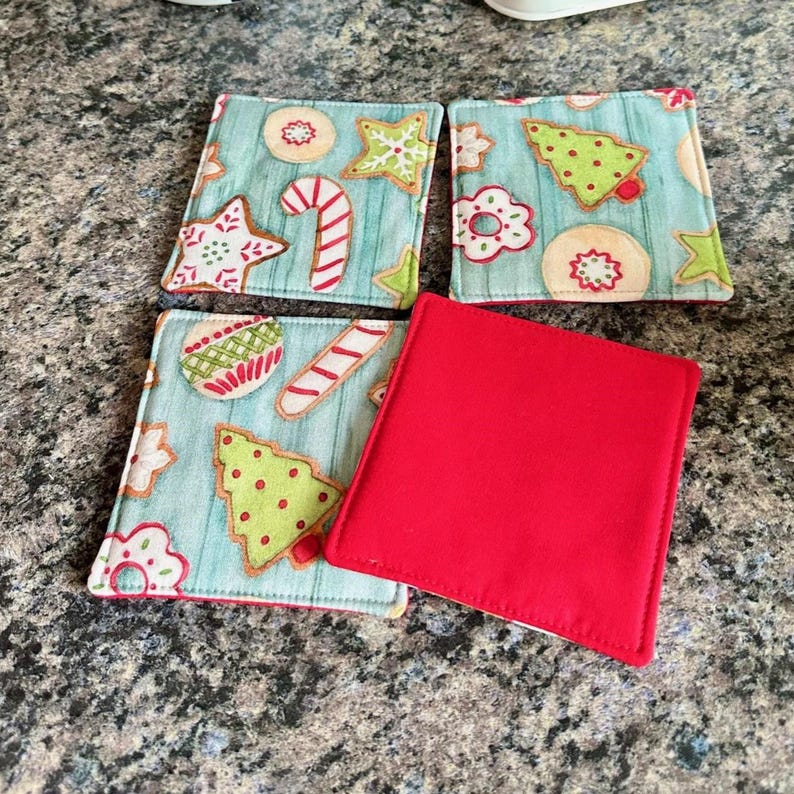 K&ouml;nnte beinhalten: Set aus vier quadratischen Untersetzern zum Thema Weihnachten. Drei Untersetzer haben einen hellblauen Hintergrund mit Lebkuchen-Designs in Rot, Gr&uuml;n und Wei&szlig;. Ein Untersetzer ist einfarbig rot. Die Untersetzer sind wahrscheinlich aus Stoff.