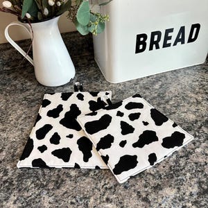 May include: Three square, black and white cow print pot holders. The pot holders are made of fabric and have a black border. A white bread box with the word "BREAD" in black lettering is in the background.