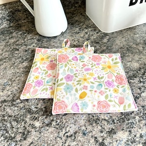 May include: Two floral print pot holders with pink, yellow, and blue flowers on a white background. The pot holders have a light pink trim.
