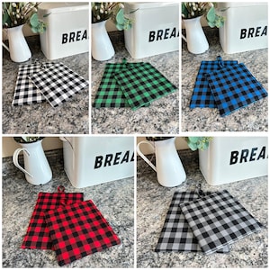 May include: A collection of buffalo check pot holders in various colors: black and white, green, blue, red, and gray. Each pot holder has a loop for hanging. A white bread box with the word "BREAD" is in the background.