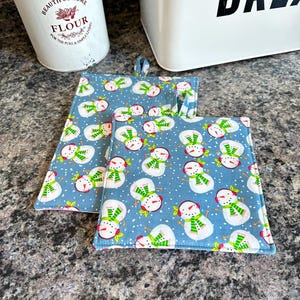 May include: Two square blue potholders with a repeating pattern of snowmen wearing green and white scarves and red hats. The potholders have a blue border and a small loop for hanging. The background is a speckled gray surface.