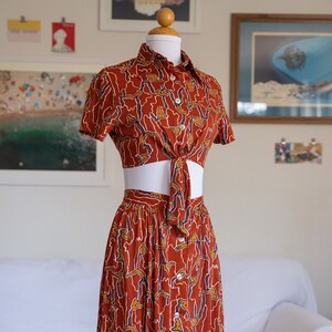 May include: A two-piece outfit with a short-sleeved crop top and a midi skirt. Both pieces are made of a brown fabric with a white and blue abstract pattern. The top has a button-up front and a tie at the waist. The skirt has a button-up front and a fitted silhouette.