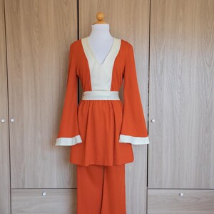 May include: A two-piece outfit in a bright orange color with a white v-neck and white trim on the sleeves. The outfit has a matching belt and wide-leg pants.