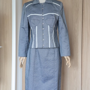 May include: A light blue suit consisting of a long-sleeved jacket and a knee-length skirt. The jacket has a collared neckline and white trim. The skirt has a straight cut. The suit is displayed on a mannequin.