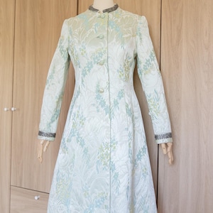 May include: A light green floral patterned coat with a mandarin collar and long sleeves. The coat features a button-down front and silver trim on the collar and cuffs. The coat is displayed on a mannequin.