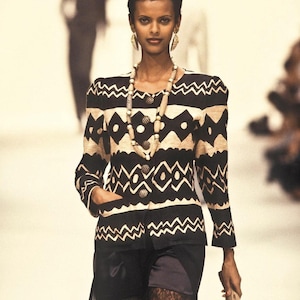 May include: A model in a black and beige patterned jacket with shoulder pads, black shorts, and a feathered headdress. The jacket features a geometric design. The model is wearing gold earrings and a beaded necklace. The image is from a fashion show.