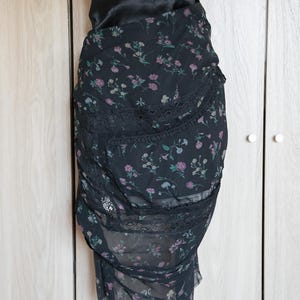 Peut inclure: Jupe noire &agrave; imprim&eacute; floral avec une taille haute et une bordure en dentelle. La jupe pr&eacute;sente un motif d&eacute;licat de petites fleurs roses, violettes et vertes sur un tissu transparent. La jupe est pr&eacute;sent&eacute;e sur un mannequin.
