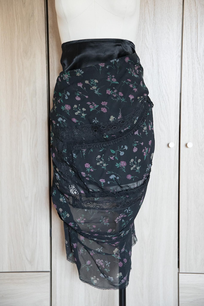 Peut inclure: Jupe noire &agrave; imprim&eacute; floral avec une taille haute et une bordure en dentelle. La jupe pr&eacute;sente un motif d&eacute;licat de petites fleurs roses, violettes et vertes sur un tissu transparent. La jupe est pr&eacute;sent&eacute;e sur un mannequin.