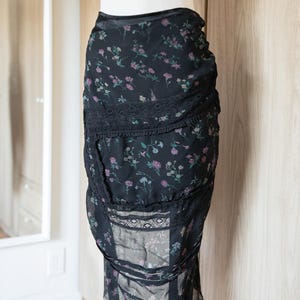 May include: Black floral print skirt with lace trim. The skirt features a delicate pattern of small pink, purple, and white flowers on a black background. The skirt is made of a sheer fabric and has a layered design.