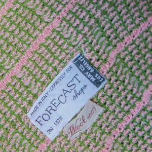 May include: A white tag with black text that reads "MADE IN ITALY - EXPRESSLY FOR FORECAST shops RN 13711" and a smaller white tag with red text that reads "Wool 100%". The tags are attached to a pink and green woven fabric.