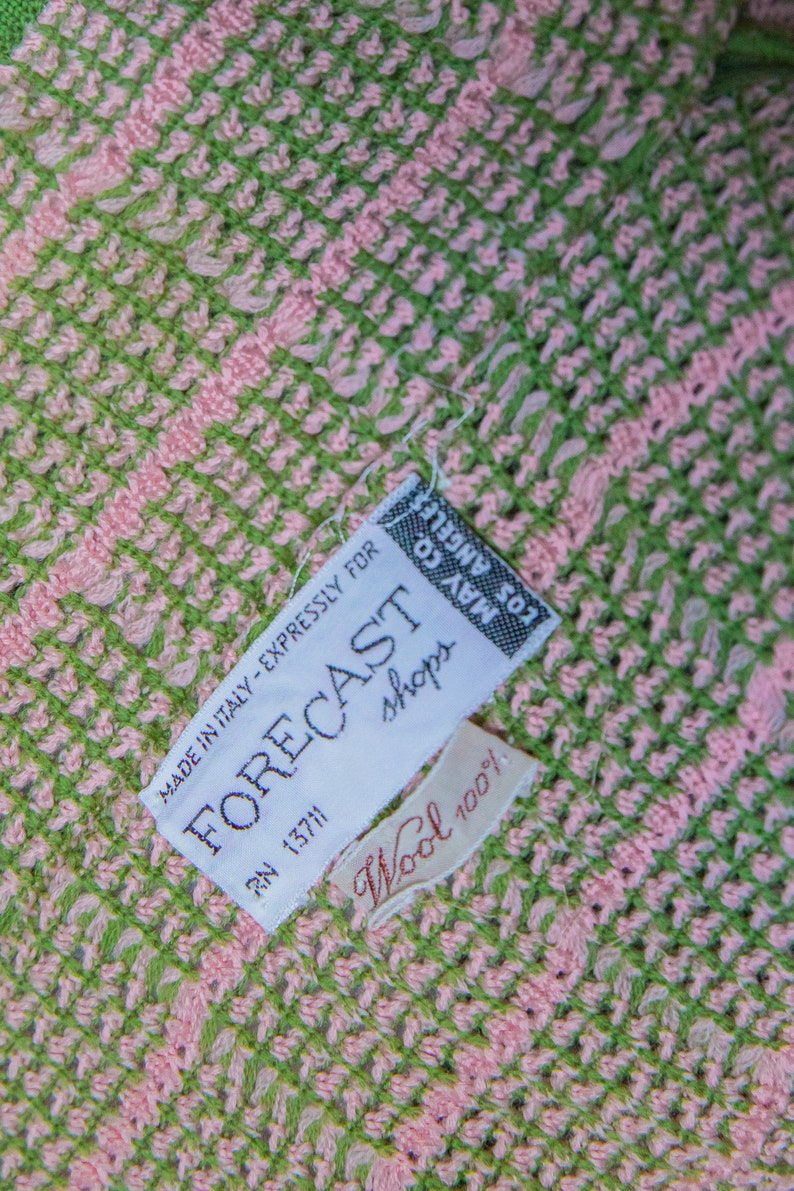May include: A white tag with black text that reads "MADE IN ITALY - EXPRESSLY FOR FORECAST shops RN 13711" and a smaller white tag with red text that reads "Wool 100%". The tags are attached to a pink and green woven fabric.
