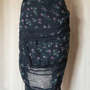 Peut inclure: Jupe noire &agrave; imprim&eacute; floral avec bordure en dentelle. La jupe pr&eacute;sente un motif d&eacute;licat de petites fleurs roses, violettes et bleues sur fond noir. La jupe a un design superpos&eacute; avec des accents de dentelle.