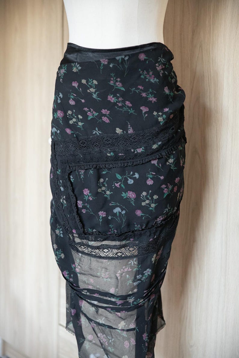 Peut inclure: Jupe noire &agrave; imprim&eacute; floral avec bordure en dentelle. La jupe pr&eacute;sente un motif d&eacute;licat de petites fleurs roses, violettes et bleues sur fond noir. La jupe a un design superpos&eacute; avec des accents de dentelle.