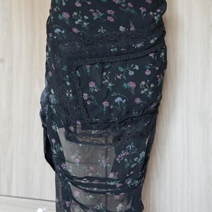 Peut inclure: Jupe noire &agrave; imprim&eacute; floral avec un design superpos&eacute;. La jupe pr&eacute;sente un fond sombre avec un motif d&eacute;licat de fleurs roses, violettes et bleues. Elle a une bordure en dentelle et un tissu transparent et fluide.