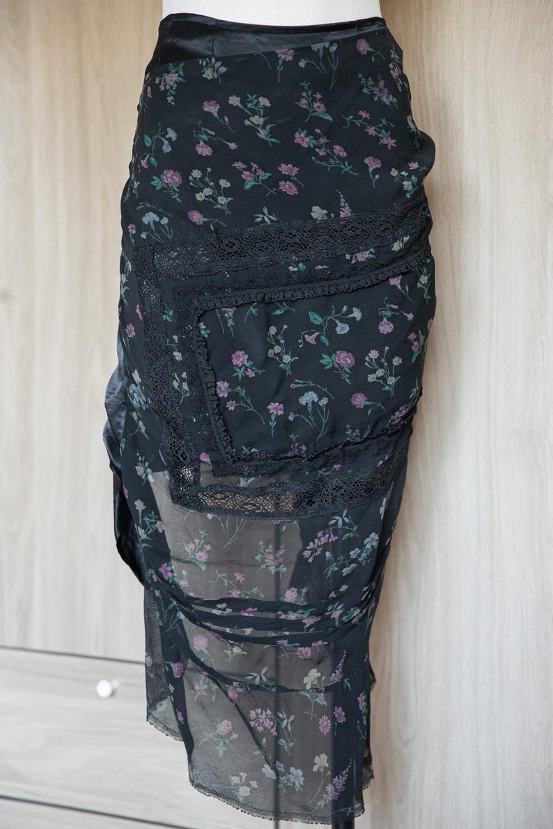 Peut inclure: Jupe noire &agrave; imprim&eacute; floral avec un design superpos&eacute;. La jupe pr&eacute;sente un fond sombre avec un motif d&eacute;licat de fleurs roses, violettes et bleues. Elle a une bordure en dentelle et un tissu transparent et fluide.